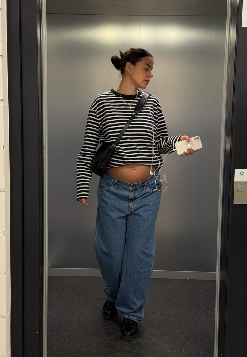 Schwarz-weiß gestreiftes langärmliges Hemd, weite blaue Jeans und schwarze Turnschuhe. Hält ein weißes Telefon mit Ohrhörern in einem Aufzug.