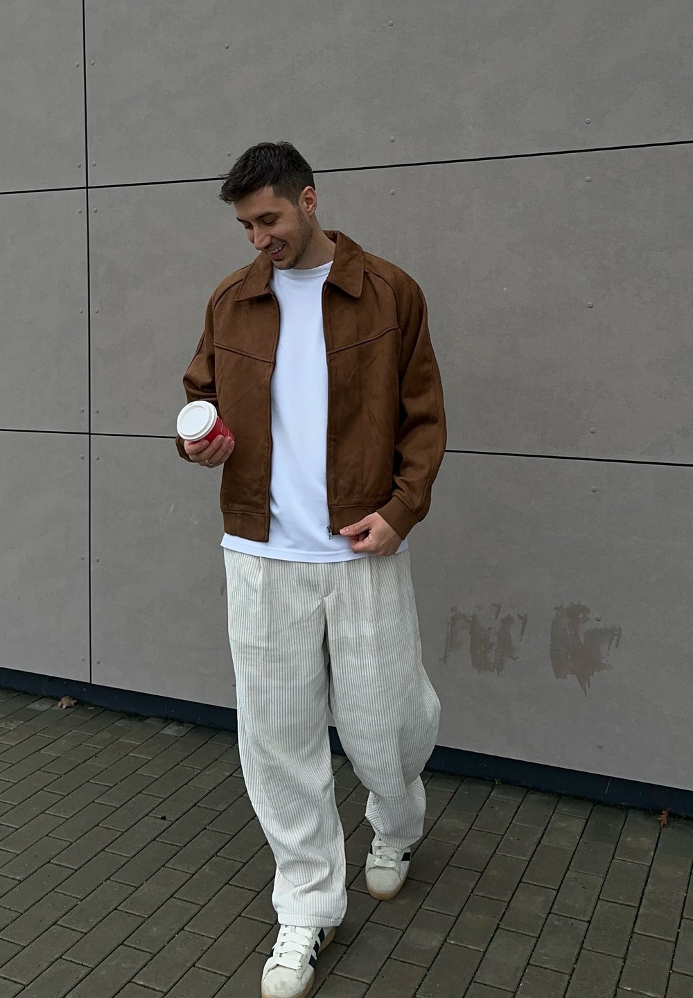 Veste en daim marron avec une chemise blanche, associée à un pantalon en velours côtelé blanc texturé. Tenant une tasse de café rose, debout sur un trottoir gris.