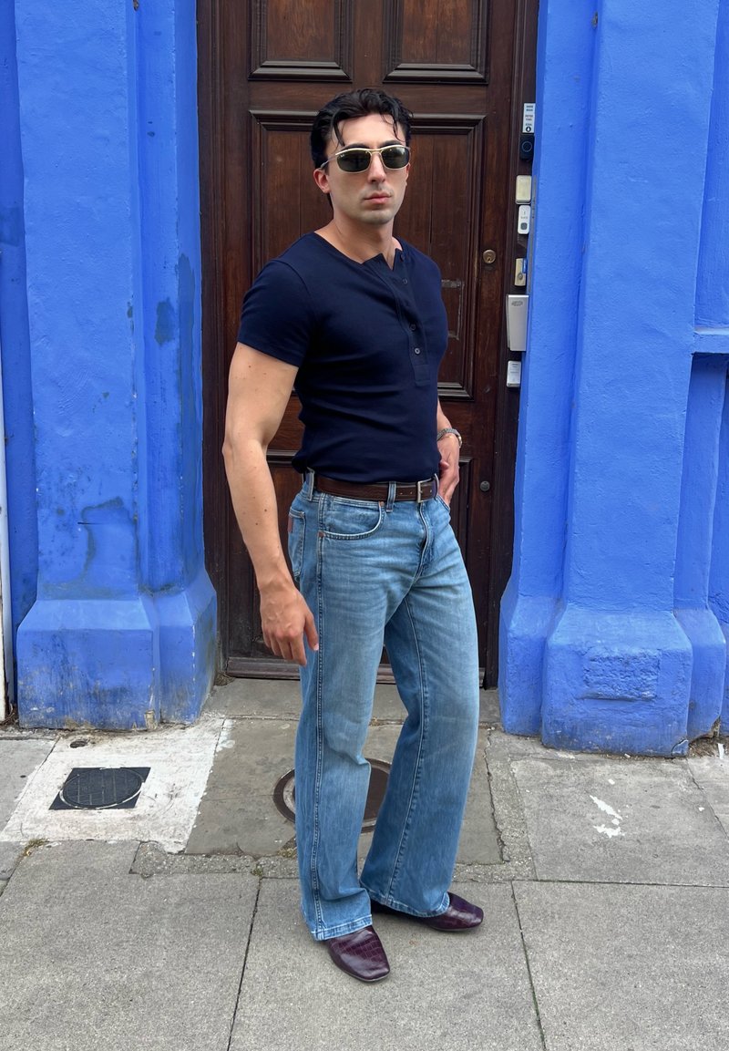 Homme portant des lunettes de soleil, une chemise noire, un jean bleu et des chaussures marron, debout devant une porte en bois encadrée par des murs bleu vif.