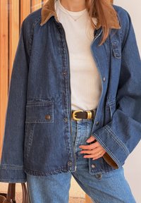 Femme portant une veste en jean bleu foncé avec un col en velours côtelé marron, un t-shirt blanc à côtes, un jean bleu, une ceinture noire à boucle dorée, et tenant un sac marron.