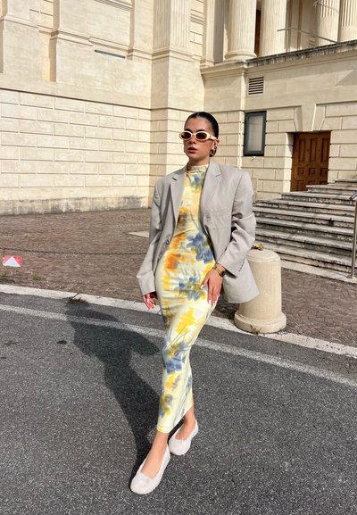 Vestido con estampado floral en amarillo, azul y naranja, con cuello alto; combinado con una chaqueta oversize gris claro y zapatos blancos peludos.