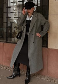 Šedý kabát z vlnenej zmesi s uvoľneným strihom, dvojradovým zapínaním a veľkými vreckami, kombinovaný s čiernymi šatami a čiernymi špicatými čižmami.