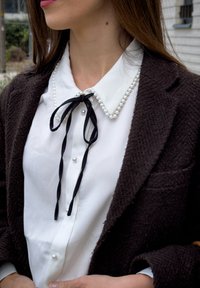 Femme portant un chemisier blanc avec des boutons en perle et un col bordé de perles, noué avec un ruban noir, sous un blazer texturé foncé en extérieur.
