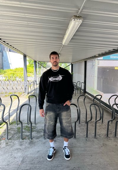 Sudadera negra con estampado gráfico, pantalones cortos de mezclilla negra desgastados, calcetines blancos y zapatillas negras. Suelo de hormigón y aparcabicis de metal en el fondo.