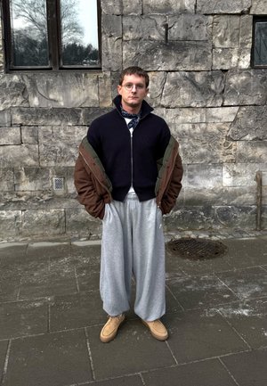 Persona vestida con un suéter azul marino con cremallera, una chaqueta bomber marrón, pantalones de chándal grises de pierna ancha y zapatillas color beige frente a una pared de piedra.