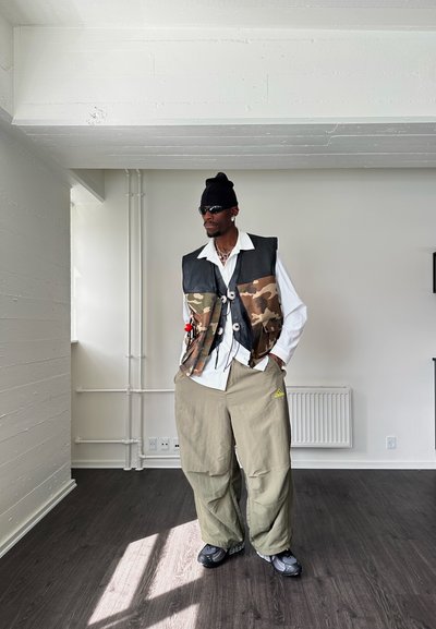 Chaleco con patrón de camuflaje y detalles en cuero negro, camisa blanca de gran tamaño y pantalones holgados en color verde oliva. Un gorro negro y gafas de sol completan el look.