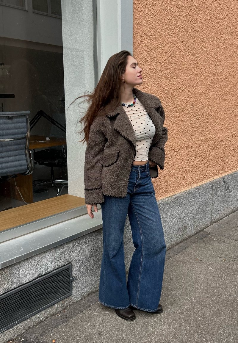 Casaco castanho fofo com gola larga, top curto bege com bolinhas, e jeans azuis com bainha ampla. Em pé contra uma parede laranja texturizada.