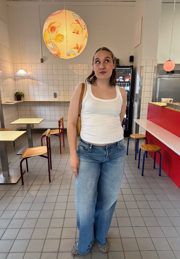 Frau in weißem Tanktop und Jeans steht in einem gefliesten Café mit einer bunten runden Lampe darüber und Tischen mit Stühlen im Hintergrund.