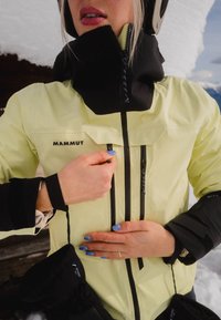 Personne en train de zipper une veste Mammut jaune et noire à l'extérieur, avec des ongles à motifs bleus et un arrière-plan enneigé.
