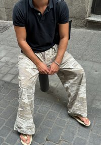 Világos színű terepmintás cargó nadrág oldalzsebekkel, fekete polóinggel és papuccsal, egy járdán ülve.