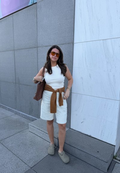 Mujer con camiseta blanca sin mangas y pantalones cortos, con jersey marrón atado a la cintura, bolso marrón, gafas de sol, de pie frente a una pared con azulejos grises y blancos.