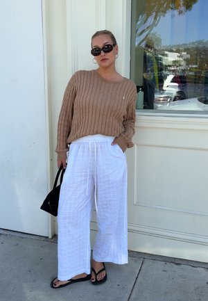 Mujer con suéter marrón de punto trenzado, pantalones blancos de pierna ancha, sandalias negras y gafas de sol, de pie contra una pared y ventana blancas.