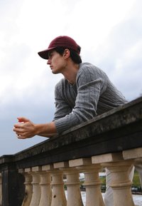 Pull gris en maille torsadée, casquette bordeaux, appuyé sur une balustrade en pierre. Mains posées sur la rambarde, arrière-plan nuageux, couleurs et textures naturelles.