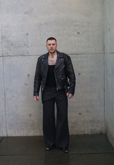 Hombre con pelo corto y tatuajes visibles en el pecho que lleva una chaqueta de cuero negra, pantalones anchos a rayas y botas, de pie contra una pared de concreto gris.