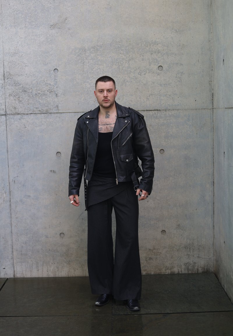 Muž s krátkymi vlasmi a viditeľnými tetovaniami na hrudi má na sebe čiernu koženú bundu, široké pruhované nohavice a topánky, stojí pri sivej betónovej stene.