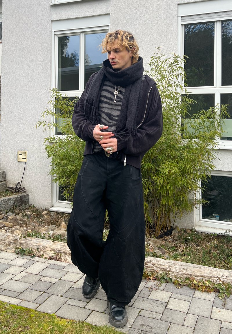 Ung person med krøllete blondt hår som har på seg svart skjerf, stripete genser, vide svarte bukser og ringer, står utendørs nær et hus med planter.