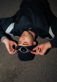 Sluneční brýle se stříbrným rámem a tmavými čočkami, které nosí osoba v černé čepici a černém sportovním tričku, ležící na texturovaném povrchu.