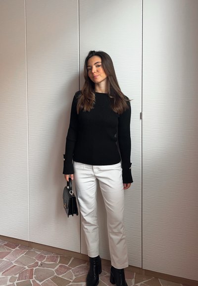 Jersey de punto negro con cuello de pico y mangas largas, pantalones blancos de pierna recta y botas negras a la altura del tobillo. Sosteniendo un pequeño bolso negro. Fondo neutro.
