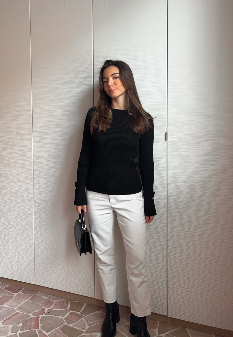 Čierny rebrovaný pletený sveter s dlhými rukávmi, biele rovné nohavice a čierne členkové čižmy. Drží malú čiernu kabelku. Neutrálne pozadie.
