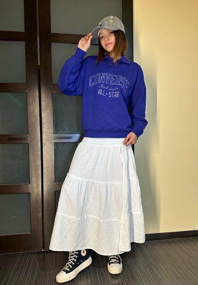 Sudadera morada con el logo "Converse All Star", falda larga blanca de capas, gorra gris con adornos y zapatillas negras de caña alta.