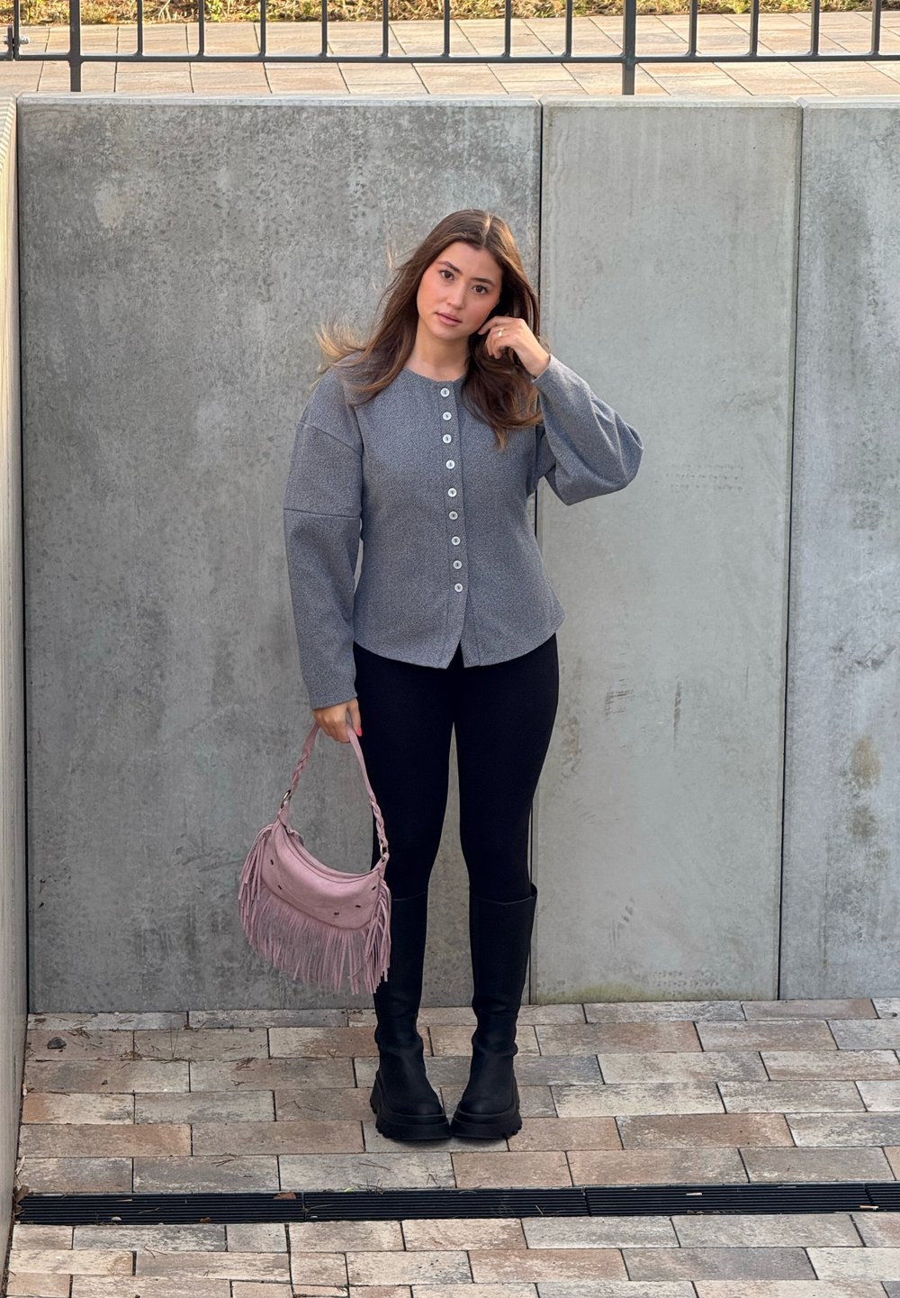 Veste boutonnée grise texturée avec manches larges, leggings noirs, bottes noires montant jusqu'aux genoux, et sac à main rose à franges.