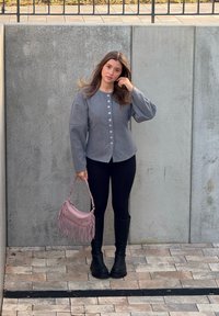 Veste boutonnée grise texturée avec manches larges, leggings noirs, bottes noires montant jusqu'aux genoux, et sac à main rose à franges.
