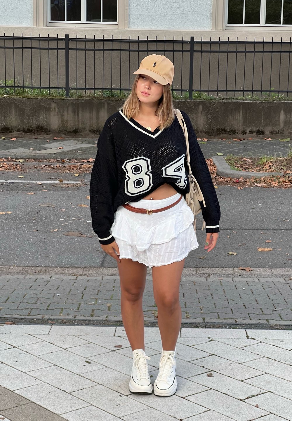 Μαύρο oversized πουλόβερ με λευκές λεπτομέρειες και τον αριθμό 84, συνδυασμένο με λευκή φούστα με φουσκωτό τελείωμα. Φοράει μπεζ καπέλο και λευκά sneakers ψηλά.