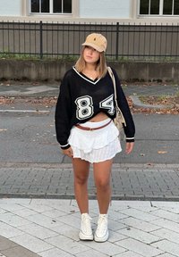 Μαύρο oversized πουλόβερ με λευκές λεπτομέρειες και τον αριθμό 84, συνδυασμένο με λευκή φούστα με φουσκωτό τελείωμα. Φοράει μπεζ καπέλο και λευκά sneakers ψηλά.