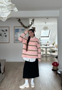 Pull oversize rayé rose et beige avec un tricot texturé, porté par-dessus une chemise blanche à col. Associé à une jupe noire et des chaussures blanches épaisses.