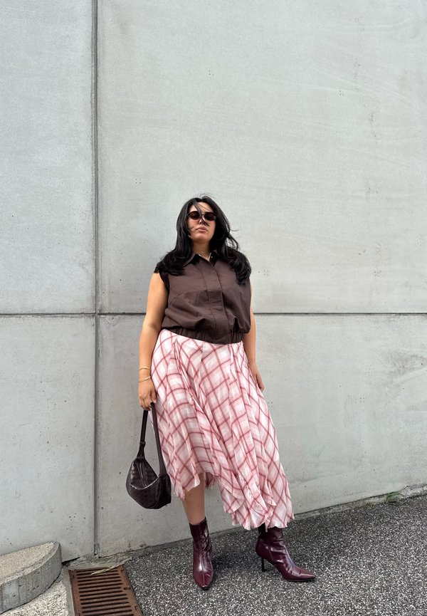 Mujer con top marrón sin mangas, falda rosa a cuadros, botas burdeos, gafas de sol y sosteniendo un bolso marrón frente a una pared gris.
