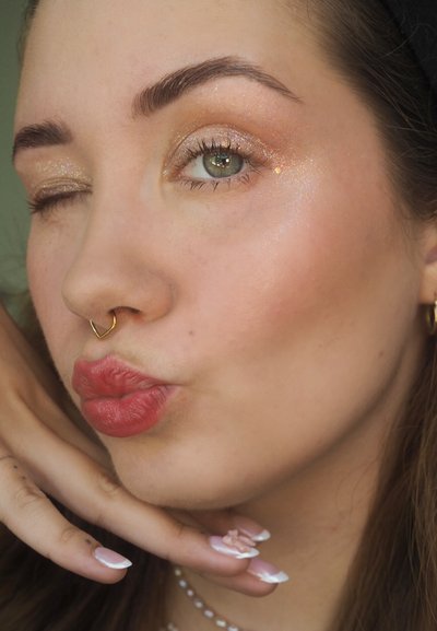 Primer plano de un rostro con un brillo dorado en los párpados, un aro septal dorado, y labios de un suave color rosa. Presenta pestañas largas y un fondo neutro.