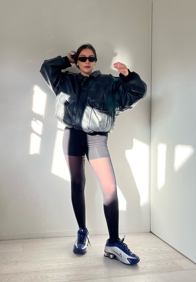 Chaqueta oversized de cuero negro con bolsillos frontales, pantalones cortos negros y zapatillas deportivas azules. El modelo lleva gafas de sol oscuras en un espacio bien iluminado.