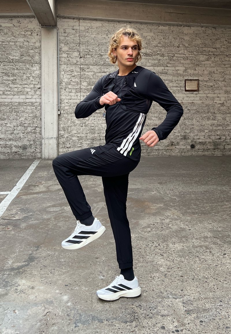 Person in black sportswear and white trainers raises one knee, posing mid-exercise in an indoor concrete space with a brick wall.