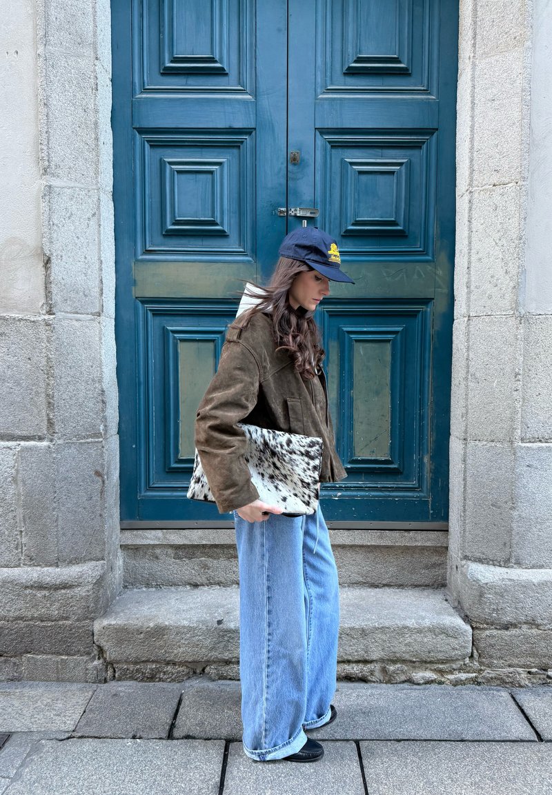 Donna con giacca marrone, cappellino blu e jeans a gamba larga che sta accanto a una porta blu chiusa, tenendo in mano una borsa a tracolla maculata.
