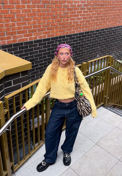 Cárdigan corto amarillo y esponjoso con botones, jeans oscuros de tiro alto y amplios, y zapatos negros con acentos plateados. Bolso de estampado de cebra y pañuelo rosa en la cabeza.