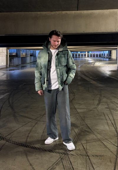 Chaqueta verde acolchada con capucha sobre una camisa blanca, combinada con pantalones grises holgados y zapatillas blancas, en un garaje con poca luz.