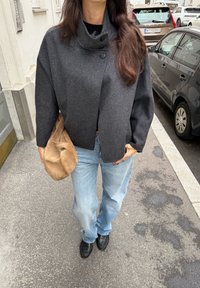 Charcoalgrijze, oversized wolblend jas met hoge kraag en knoopsluiting aan de voorkant; lichtblauwe jeans; houdt een tan suède tas vast; zwarte schoenen.