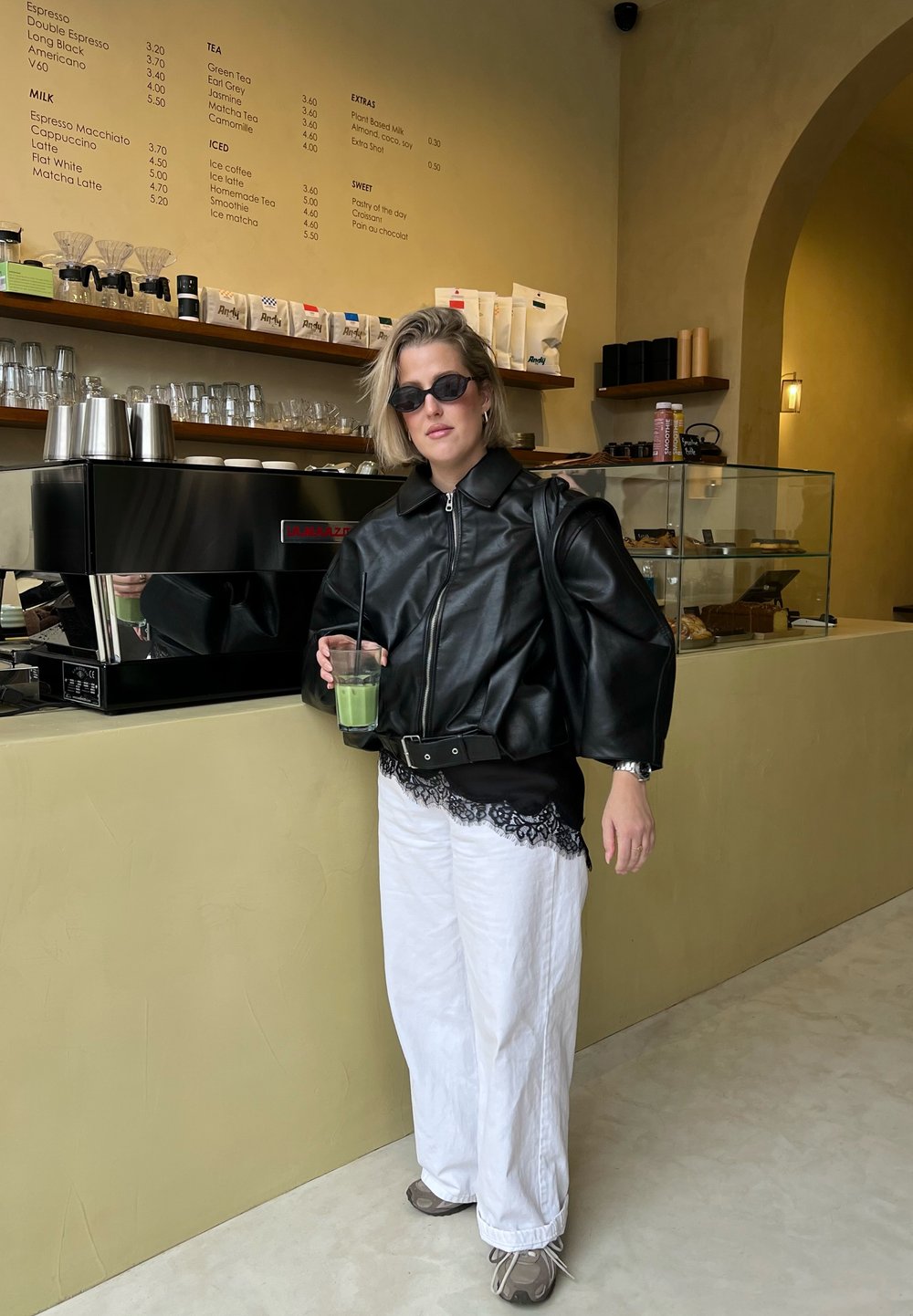 Veste en cuir noire avec manches surdimensionnées, chemise blanche à dentelle, pantalons blancs amples et baskets grises. Tenant une boisson verte.