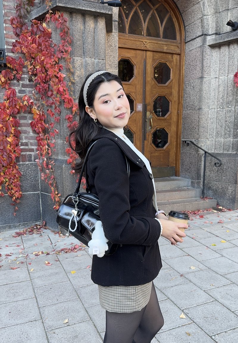 Jonge vrouw in een zwarte jas en een houndstooth rok houdt een koffiekop vast, terwijl ze staat bij een houten boogdeur en rode herfstklimop op een stenen muur.