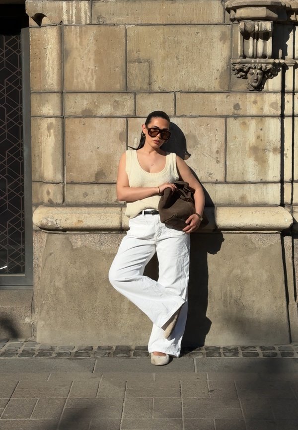Femeie purtând pantaloni albi și un top fără mâneci se sprijină de un perete de piatră ținând o geantă maro, în lumina puternică a soarelui.