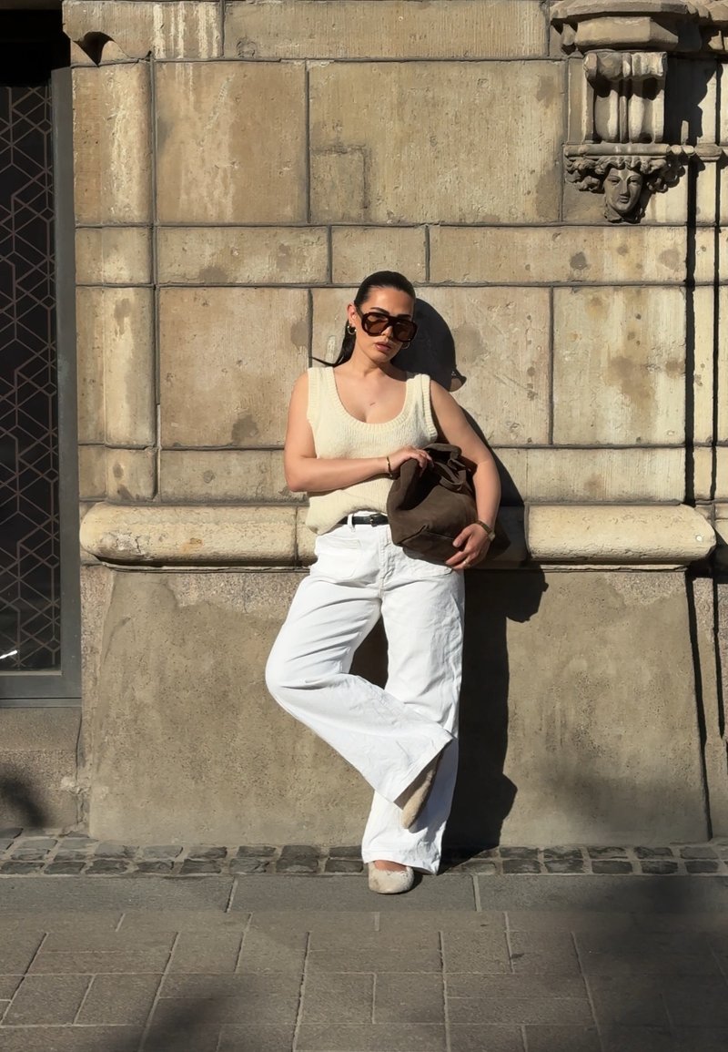 Femme portant un pantalon blanc et un haut sans manches, appuyée contre un mur de pierre, tenant un sac marron sous un soleil éclatant.