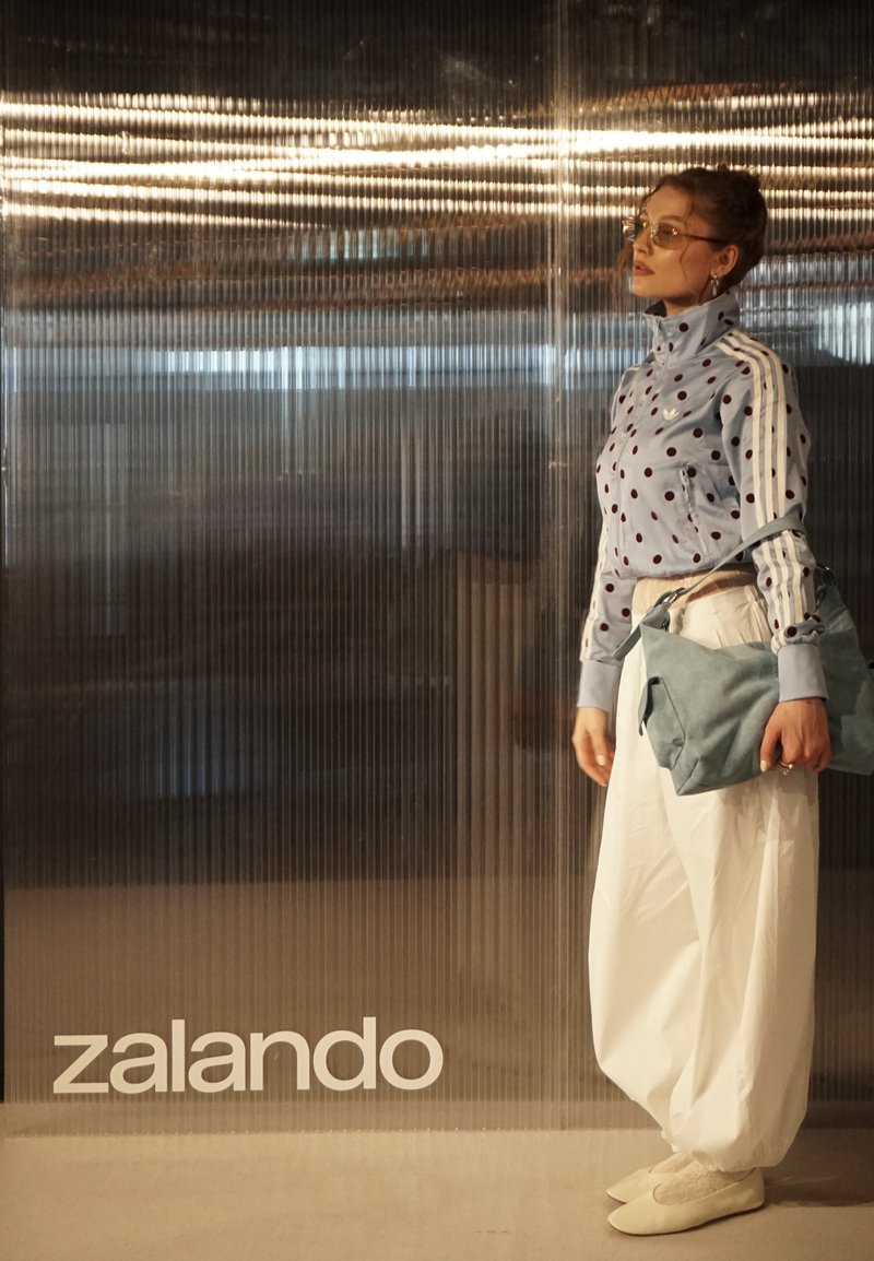 Kobieta w kurtce w groszki i białych szerokich spodniach, niosąca niebieską torbę, stojąca przy ryflowanej szklanej ścianie z napisem "zalando".