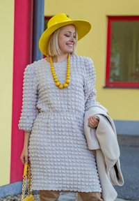 Γκρι φόρεμα με υφή, με φουσκωτά μανίκια, σφιχτή μέση και κίτρινο κομπολόι. Συμπληρώνεται από ένα κίτρινο καπέλο με φαρδύ γείσο και κρεμ παλτό.