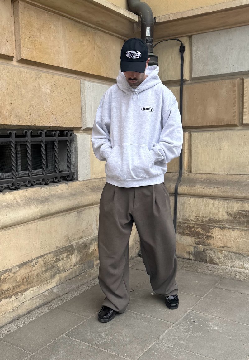 Homme portant un sweat à capuche gris OBEY, casquette noire, pantalon brun large et chaussures noires, debout contre un mur en pierre sur un trottoir en ville.