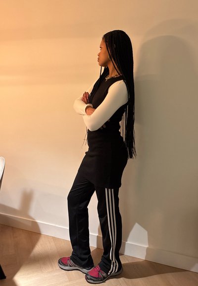 Top sin mangas negro sobre una camiseta de mangas largas blanca, combinado con pantalones negros con rayas blancas y zapatillas oscuras rojas. Trenzas largas.