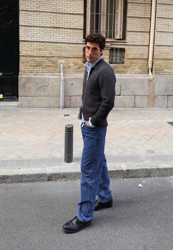 Maglione grigio scuro con cerniera sopra una camicia azzurro chiaro, jeans blu e scarpe nere; in piedi su una strada con un muro di mattoni e un palo nelle vicinanze.