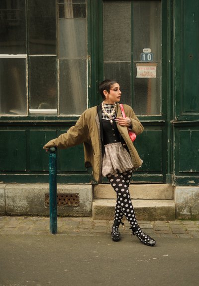 Chaqueta verde de gran tamaño, blusa a cuadros en blanco y negro, falda con capas, medias con puntos y zapatos estampados frente a una pared verde.