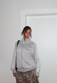 Nainen, jolla on kihara tukka, pukeutuneena hopeanväriseen Adidas-treeni-takkiin ja naamiohousuihin, seisoo sisällä valkoista seinää ja ovea vasten.