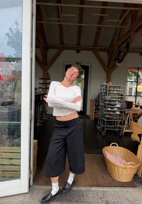Young person with short curly hair, white long sleeve shirt, and black wide pants smiling with arms crossed at cafe entrance.