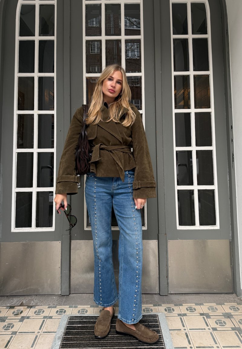 Jeune femme aux longs cheveux blonds portant une veste marron ceinturée, un jean bleu à clous et des mocassins marron, debout devant des portes grises.